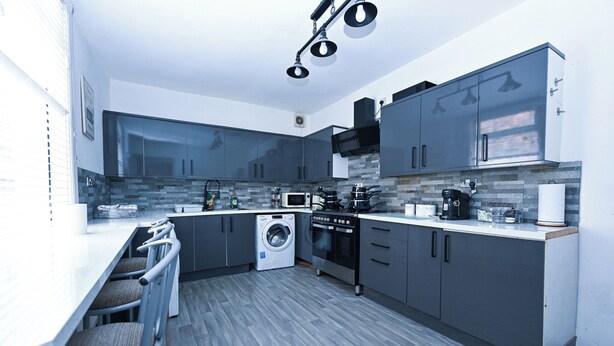 Une cuisine moderne avec des meubles gris, un four encastré, un lave-linge et un coin bar avec des tabourets.