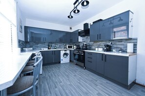 A modern kitchen with grey cabinets, a built-in oven, a washing machine, and a bar area with chairs.