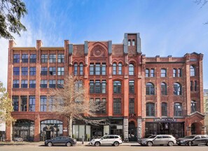 A multi-story brick building with a prominent clock tower, storefronts on the ground floor, and parked cars in front. - Historic DT Loft w/ 16ft Ceilings, Garage &97 WS! (Seattle)