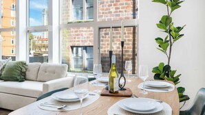 A dining area with a wooden table set for four, surrounded by large windows with a view of a brick building.