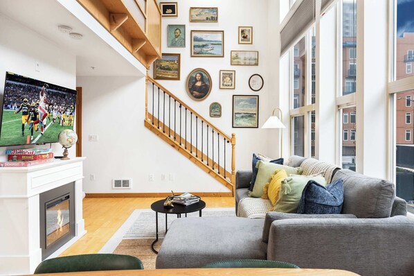 A modern living room with a fireplace, a large flat-screen TV, a staircase, and various framed artworks on the walls. - Historic DT Loft w/ 16ft Ceilings, Garage &97 WS! (Seattle)