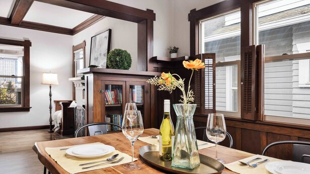 Un comedor con una mesa de madera dispuesta para dos personas, un florero con flores y una estantería al fondo.