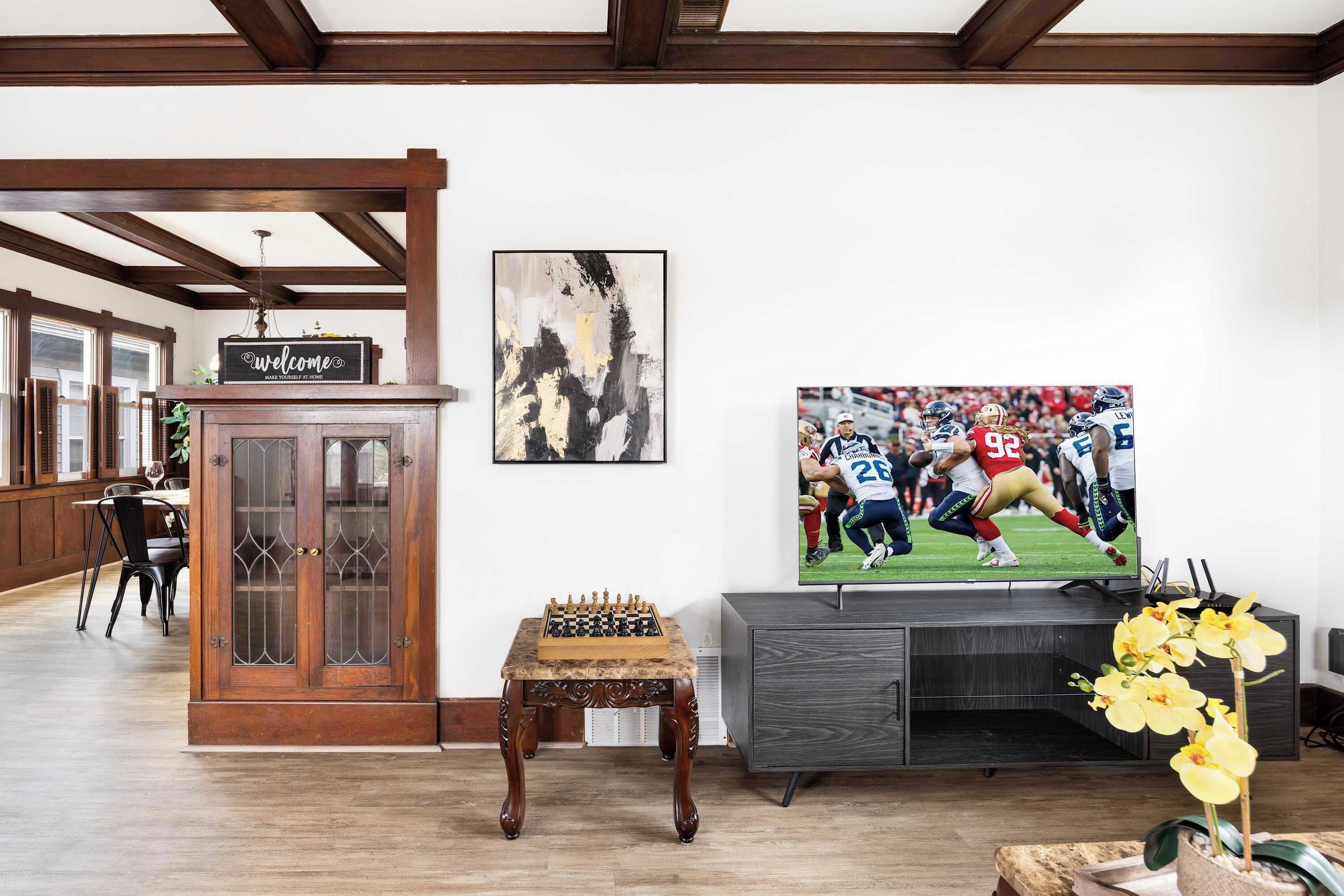 Uma sala de estar com um armário de madeira, uma mesa de xadrez, uma TV exibindo uma partida de futebol e um letreiro de boas-vindas.