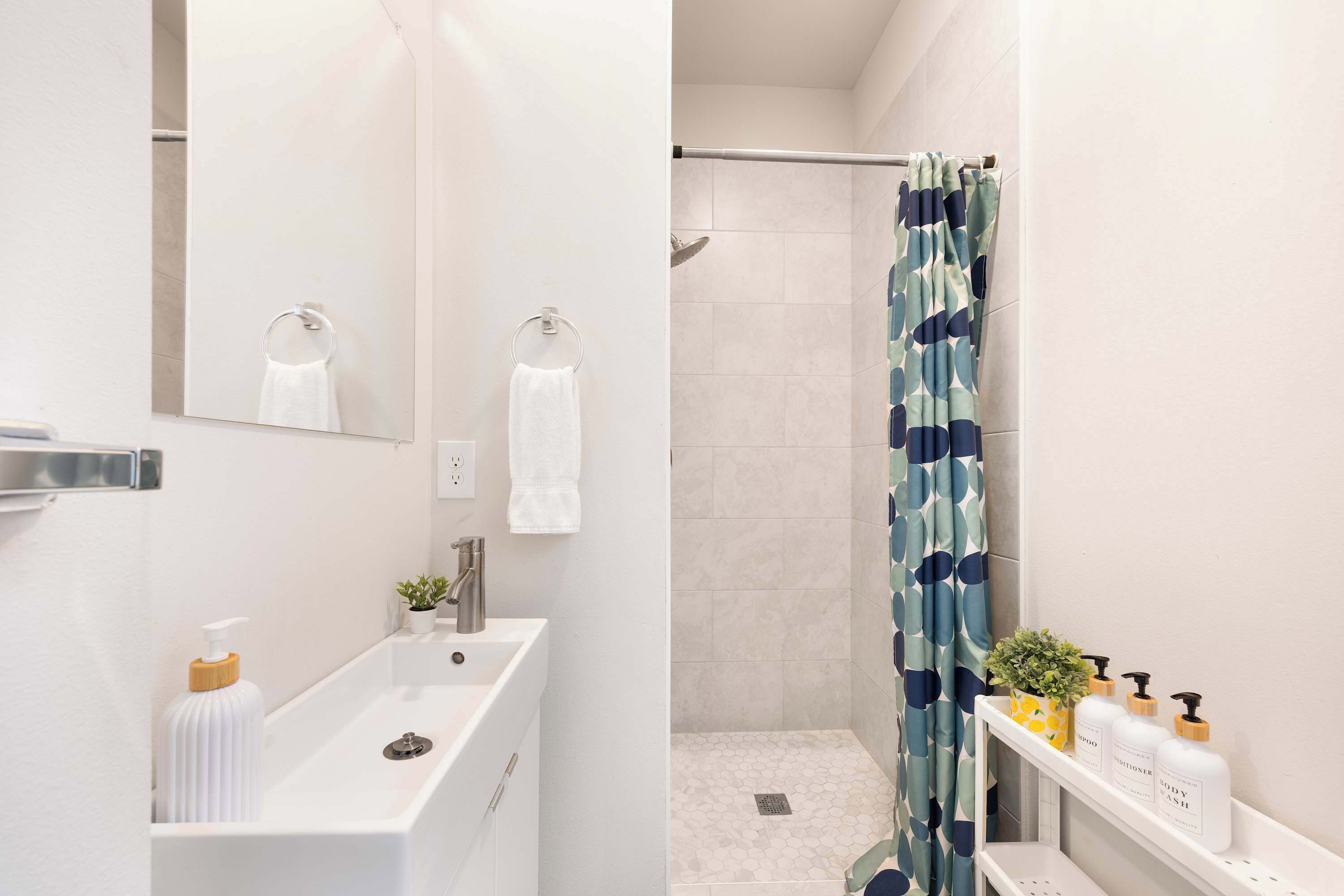 Et moderne badeværelse med en hvid vask, et spejl, en bruser med et blåt mønstret gardin og toiletartikler på en hylde.