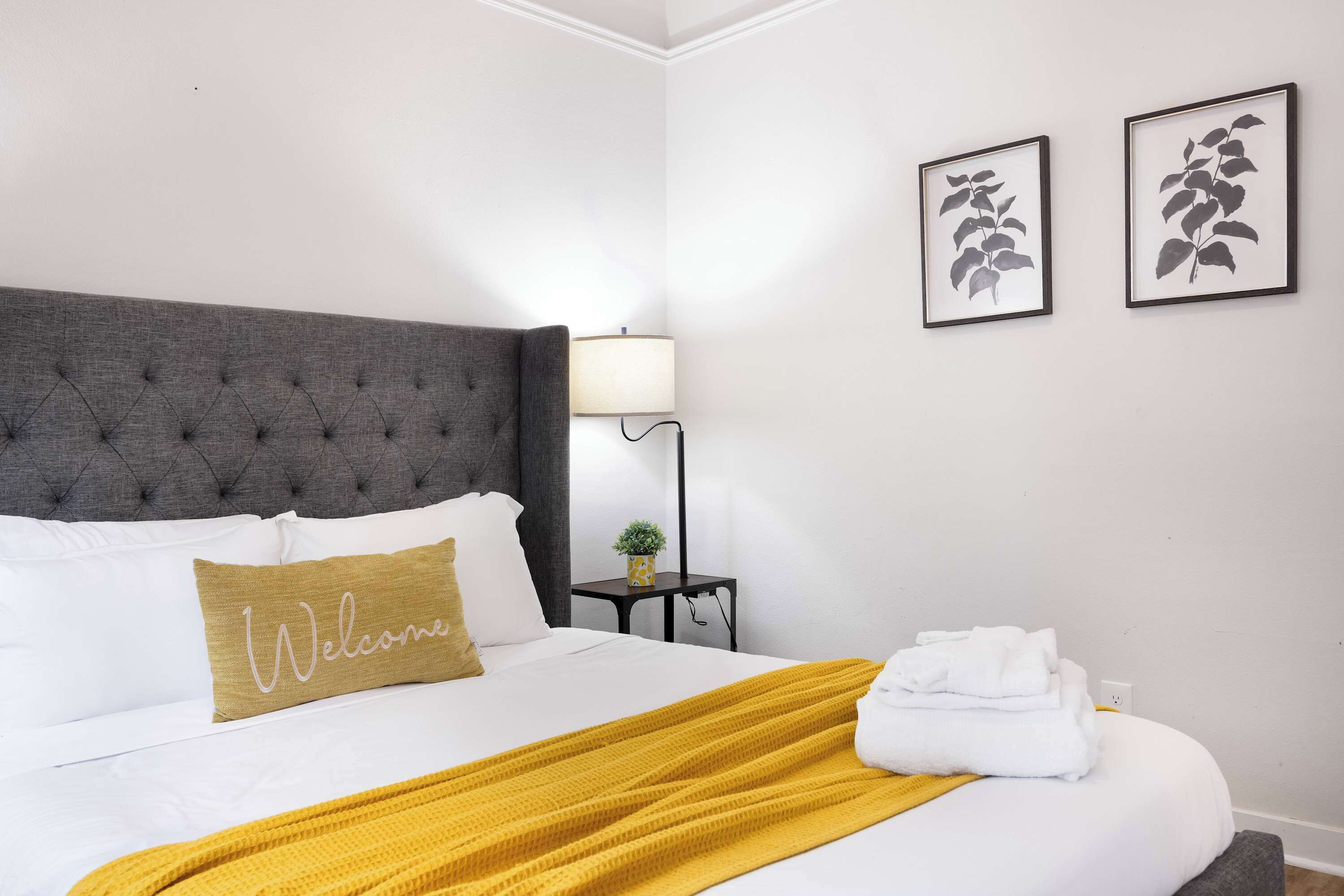 A neatly made bed with a 'Welcome' pillow, a grey tufted headboard, a small bedside table with a lamp, and two framed leaf artworks on the wall.