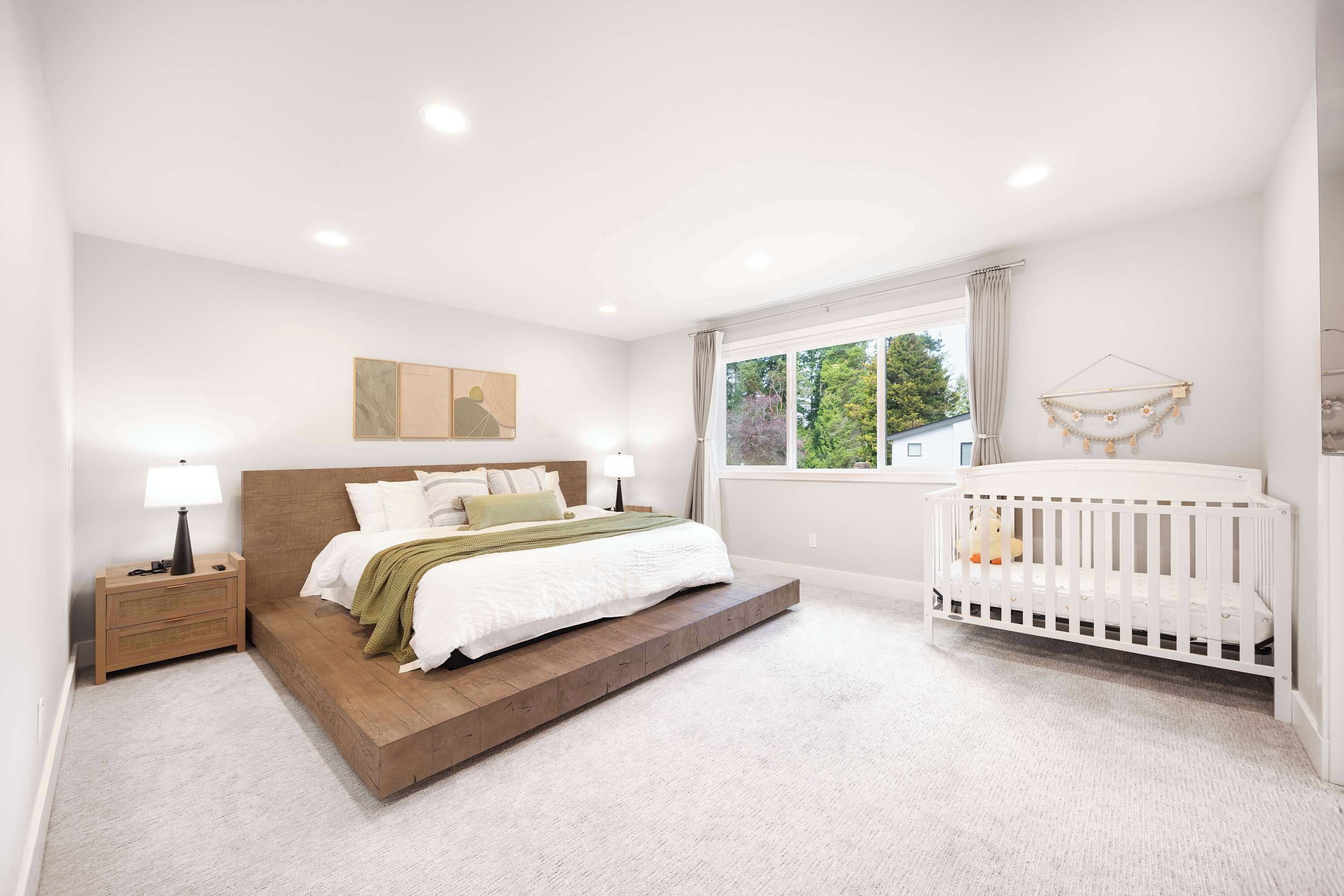A bedroom with a bed, nightstand, crib, and a window with a view of trees.