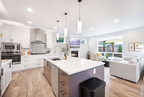 A modern kitchen with white cabinetry, a large island, stainless steel appliances, pendant lighting, and a fireplace.
