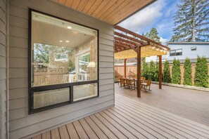 Un patio cubierto con cubierta de madera, una pérgola con luces colgantes y vistas a un jardín trasero con cerco de madera y follaje.