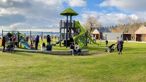 Un parque infantil con toboganes y estructuras para escalar, rodeado de un campo cubierto de césped y edificios al fondo.