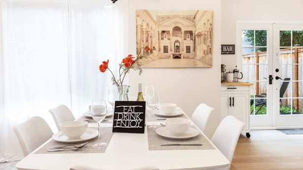 Un comedor con una mesa dispuesta para cuatro, una araña, un cuadro de un edificio y un letrero que dice "EAT DRINK ENJOY".
