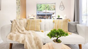 Una sala de estar moderna con un conjunto de sofás, una planta en maceta y vistas de una casa a través de un amplio ventanal.