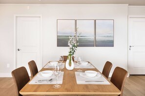 Ein Esszimmer mit einem Holztisch, gedeckt für sechs Personen, weißen Stühlen und einer Blumenvase. Darüber an der Wand befinden sich drei gerahmte Bilder.