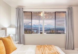 A hotel room with a large window offering a city view, a bed with white bedding and a beige throw, and a nightstand with a lamp. - Luxury Bellevue 2B Basement w, Stunning View (Bellevue)
