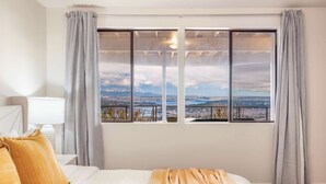 A hotel room with a large window offering a city view, a bed with white bedding and a beige throw, and a nightstand with a lamp. - Luxury Bellevue 2B Basement w/ 180º+ Stunning View (Bellevue)