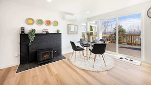 A modern living room with a fireplace, dining table, and chairs, large windows with a view of a deck.