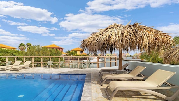 Un área de piscina con sillones de descanso y una sombrilla de paja, rodeada por una valla de madera y palmeras.