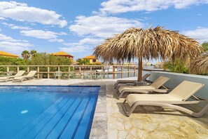 Un área de piscina con sillones de descanso y una sombrilla de paja, rodeada por una valla de madera y palmeras.