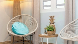 Una habitación con dos sillas de mimbre, una con cojín, y una pequeña mesa redonda con una planta y objetos decorativos.