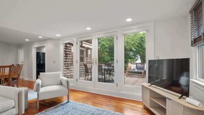 A spacious living room with a large TV, comfortable seating, and a view of the outdoors through sliding glass doors. - Rare Private Deck, Dog Friendly, Walk to Harbor! (Provincetown)