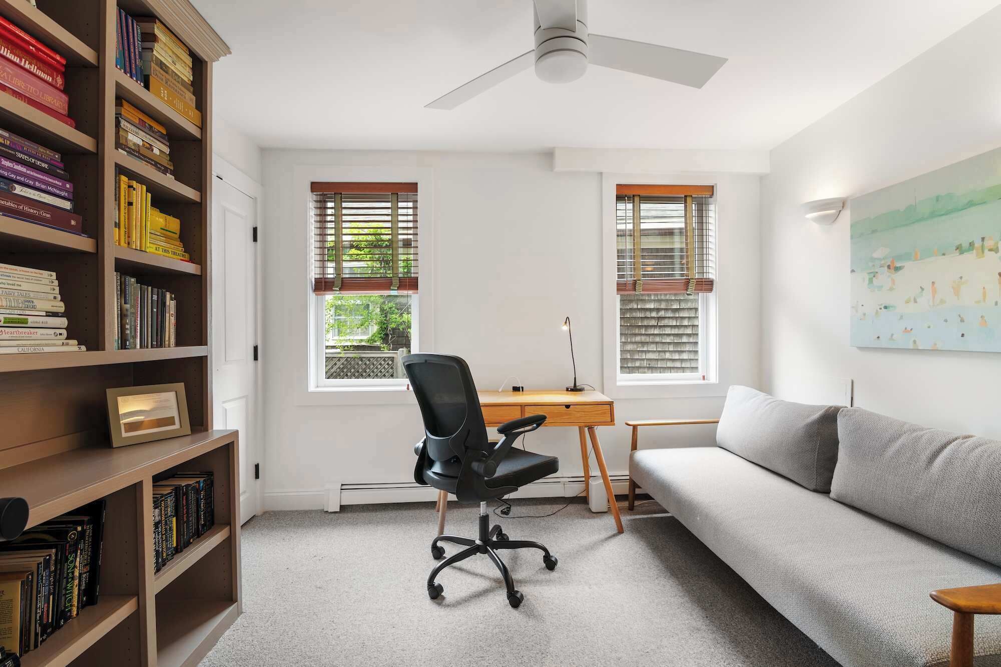 Una habitación luminosa con un escritorio, una silla de oficina negra, un sofá gris y una estantería llena de libros.
