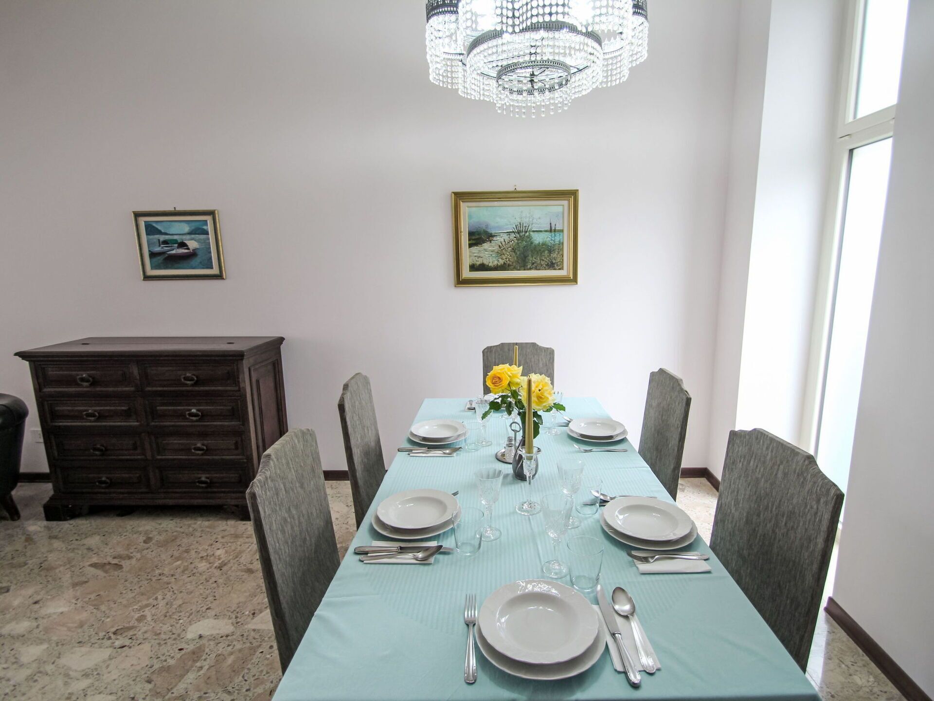 A dining room with a table set for four, a chandelier, and two framed pictures on the wall.