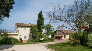 A two-story house with a gravel driveway, surrounded by lush greenery and flowering plants. - Cosy apartment for 4 people with A/C, WIFI, pool, TV, patio and pets allowed (Città Sant'angelo)