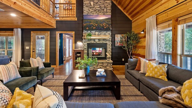 A spacious living room with a stone fireplace, wooden ceiling, and a balcony.