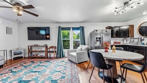 A modern living room with a flat-screen TV, a dining area with a round table, and a kitchen with dark cabinets.