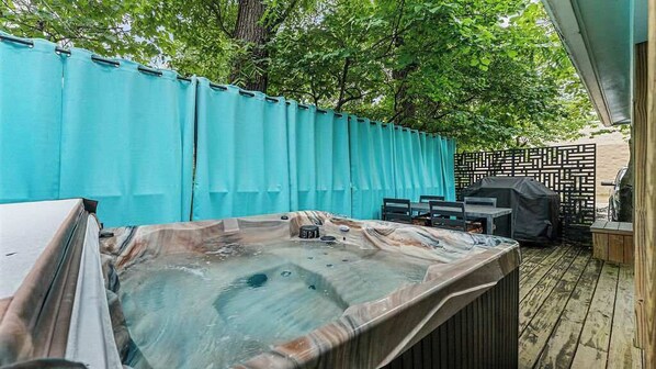 A hot tub with a wooden deck, a covered grill, and a fence.
