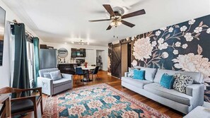 A living room with a sofa, chairs, a coffee table, and a patterned rug. There is a ceiling fan, a dining area with a table and chairs, and a kitchen in the background. - Traveling together but want space and privacy? (Eureka Springs)