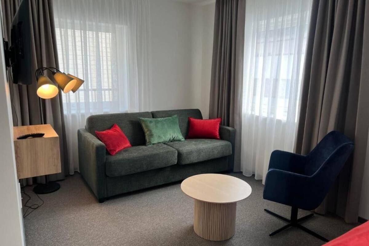Un salon moderne avec un canapé gris, des coussins rouges et verts, une table basse ronde, un fauteuil bleu et un téléviseur posé sur un meuble.