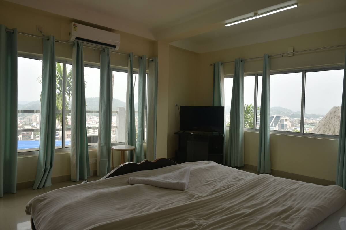 A hotel room with a bed, a television, and a view of a cityscape through large windows.