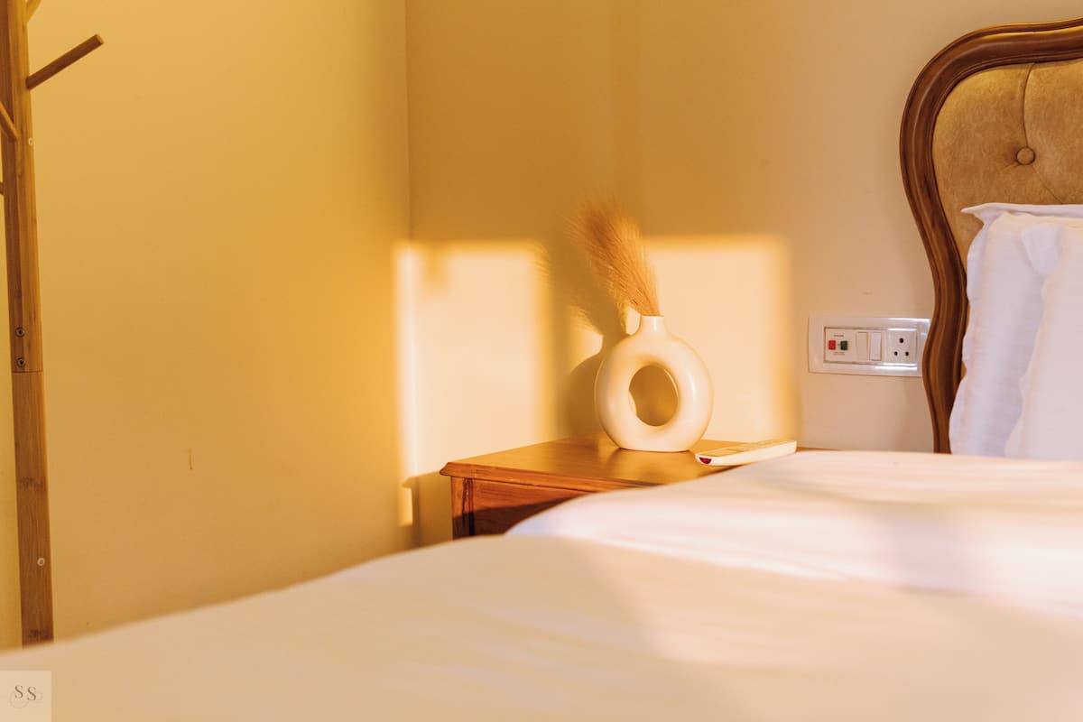 A neatly made bed with white linens, a wooden nightstand with a white vase, and a wall socket.