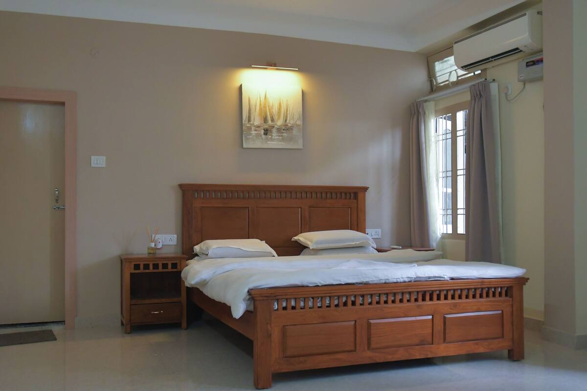 A bedroom with a wooden bed, nightstands, a wall-mounted light, an air conditioning unit, and a window with curtains.