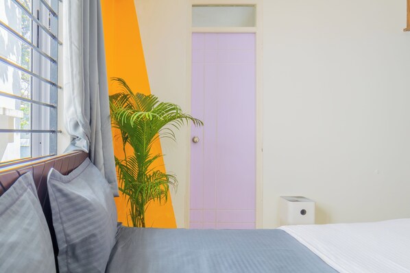 Une chambre à coucher avec un lit, une fenêtre avec des rideaux, une plante en pot et une porte avec un panneau de couleur violet clair.