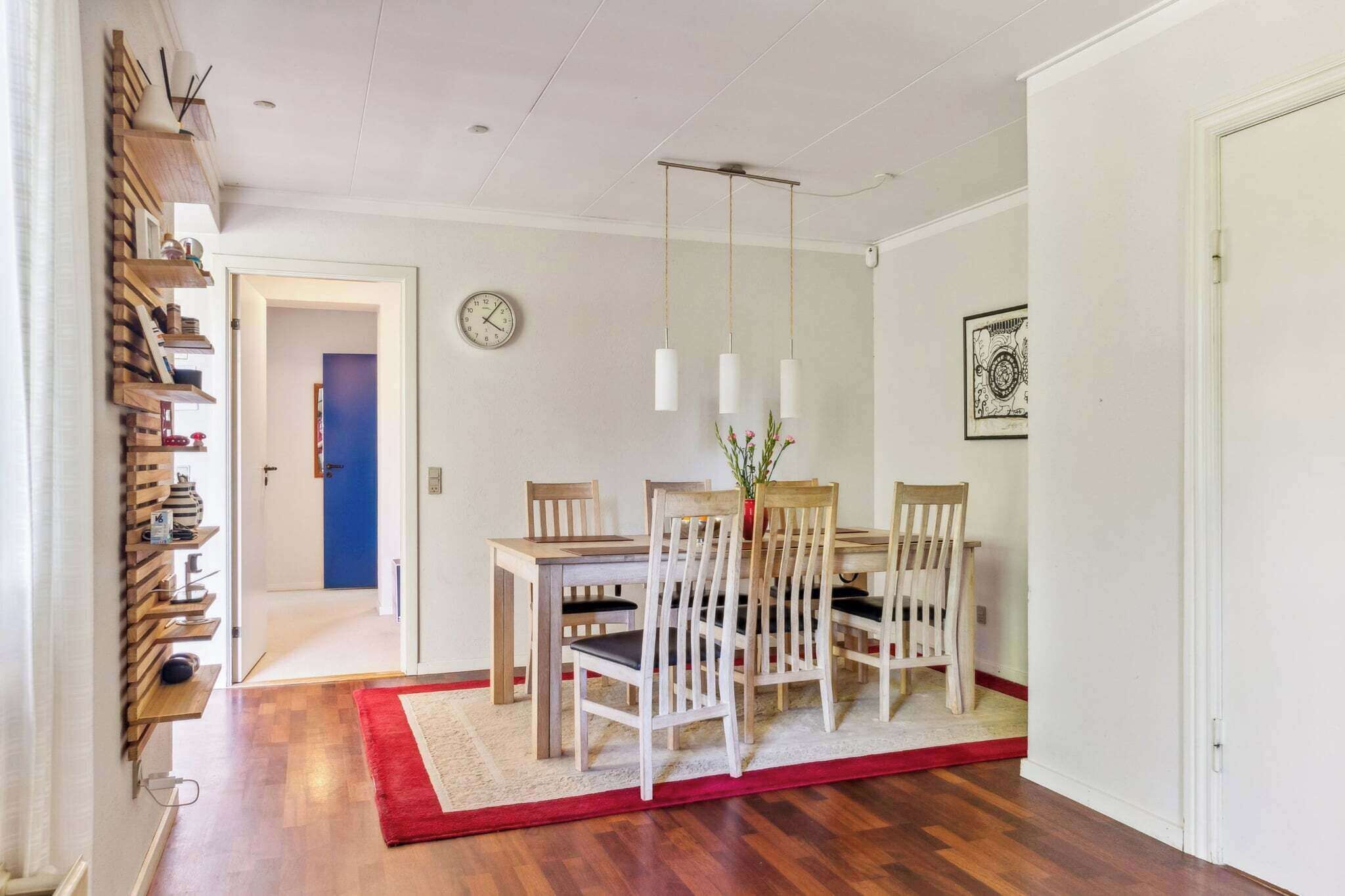 Un comedor con una mesa y sillas de madera, un tapete rojo, un reloj en la pared y una puerta que da a otra habitación.