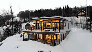 Une cabane moderne à plusieurs niveaux, dotée d’une façade en verre, située dans une forêt enneigée.