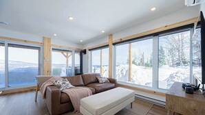 Un salon moderne avec un canapé en cuir marron, un pouf beige et un bureau en bois sur lequel se trouve un ordinateur. De grandes fenêtres offrent une vue sur un paysage enneigé.