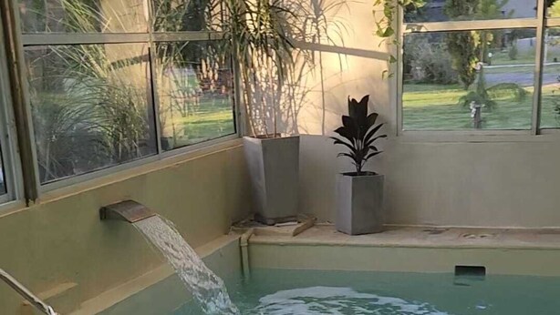 A swimming pool with a water feature, surrounded by windows and indoor plants. - Hosteria Santa Maria (Mercedes)