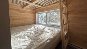A room with a bed, wooden walls, a window with a view of snow-covered trees, and a sloped ceiling with wooden beams.