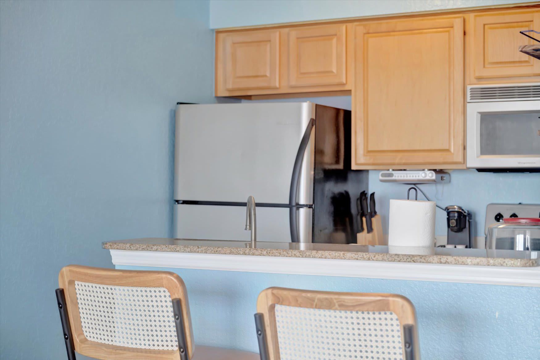 A compact kitchen with wooden cabinets, a stainless steel refrigerator, a microwave, a toaster, and two bar stools with woven seats.
