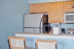 Cocina compacta con armarios de madera, un refrigerador de acero inoxidable, un microondas y una pequeña encimera con un tostador. Hay dos sillas con asientos de mimbre.