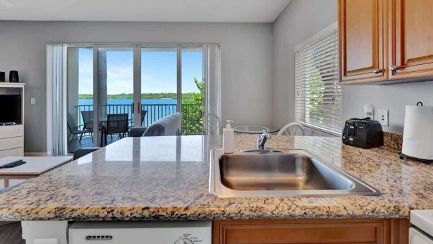 A kitchen with a granite countertop, stainless steel sink, wooden cabinets, and a view of a balcony with outdoor furniture. - Waterfront Condo w/ Private Balcony & Heated Pool (St. Petersburg)
