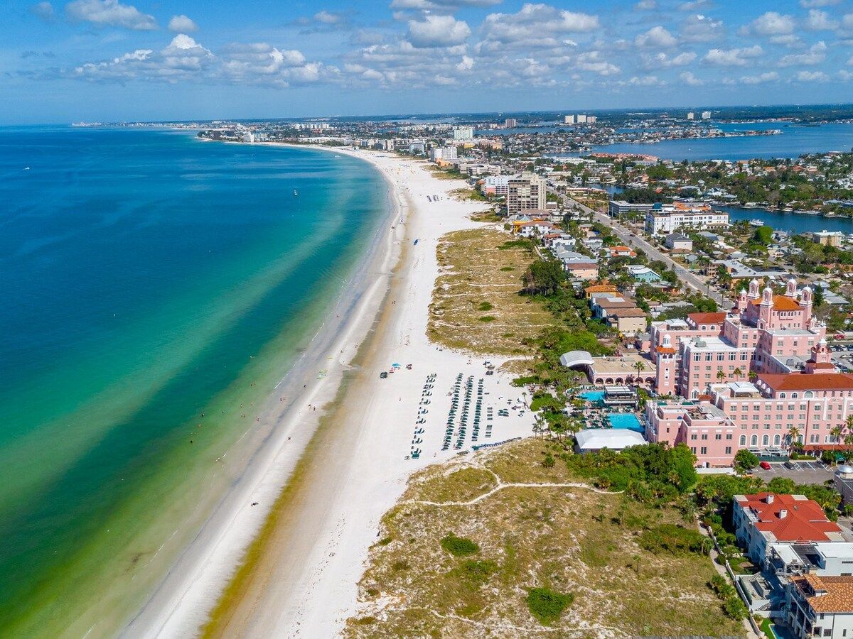 Aerial view showcasing the pristine white-sand beach stretching along turquoise Gulf waters, with the property's distinctive pink architecture and pools nestled among coastal neighborhoods.