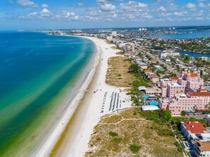 Aerial view showcasing the pristine white-sand beach stretching along turquoise Gulf waters, with the property's distinctive pink architecture and pools nestled among coastal neighborhoods. - Waterfront heated pool, dock, kayaks and bikes (St. Pete Beach)