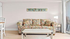 Een woonkamer met een bank met bloemenprint, een houten salontafel, een witte stoel en een bijzettafeltje met een lamp. Een bordje met de tekst 'BEACH HOUSE' hangt aan de muur.