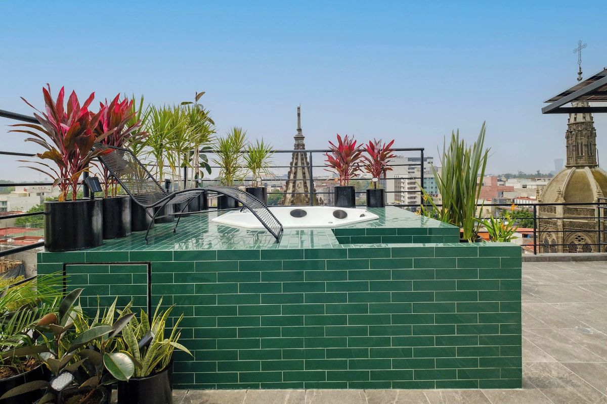 Un espace piscine aménagé sur le toit, avec un jacuzzi, entouré de plantes en pot et offrant une vue sur les bâtiments de la ville.