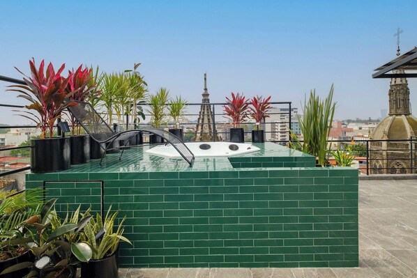 A rooftop pool area with a hot tub, surrounded by potted plants and a view of city buildings.