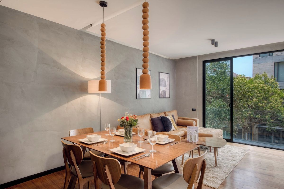 Un espace de repas moderne avec une table en bois dressée pour quatre personnes, un canapé et une grande fenêtre donnant sur un paysage verdoyant.