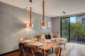 A modern dining area with a wooden table set for four, a sofa, and a large window offering a view of greenery.